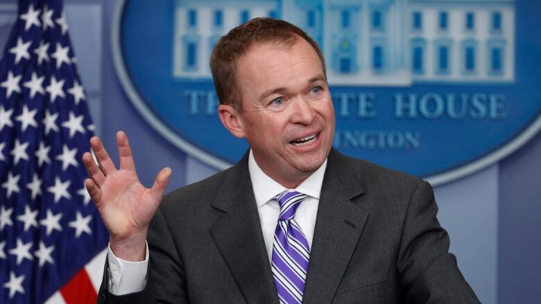 A man wearing a grey suit and blue tie holds his right hand up in a gesture while speaking at a podium. The background is a blue wall with an insignia of a building that reads: The White House, Washington.