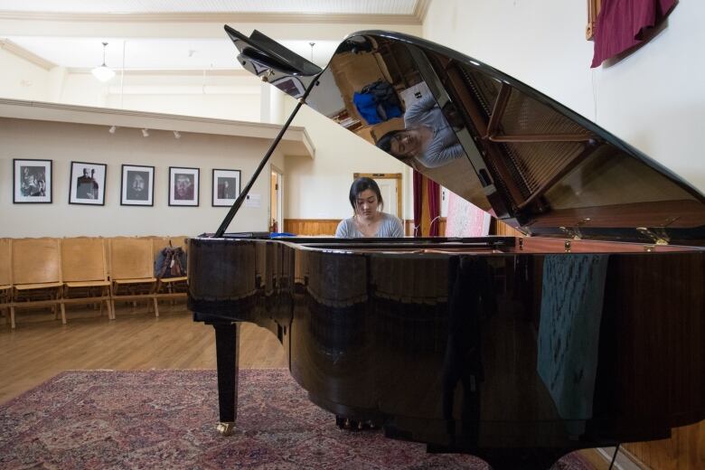 A young woman with dark hair tired back sits at a large black grand piano in an empty room