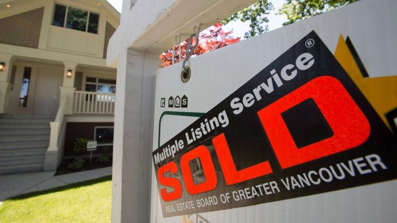 A 'for sale' sign stands on a lawn in front of a detached house. A sticker across it says 'sold.'