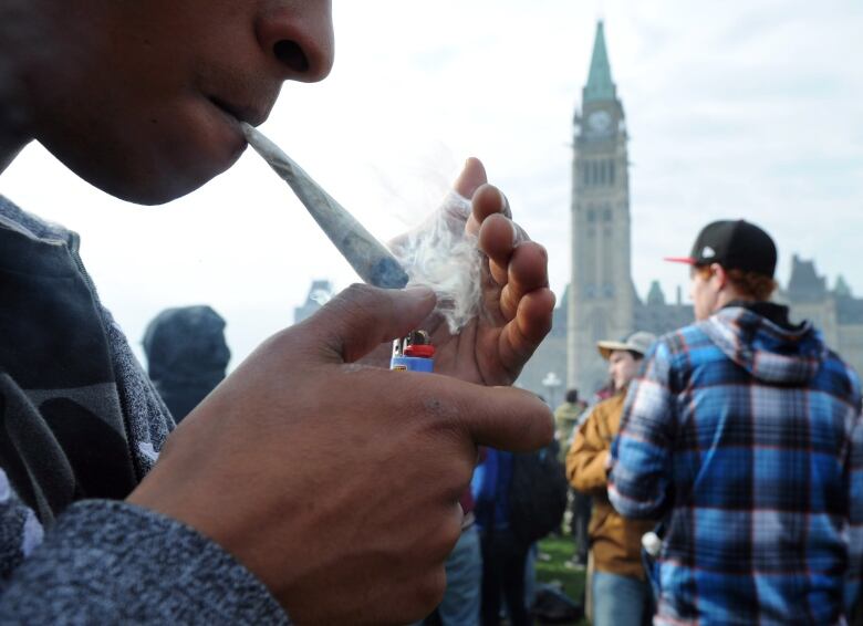 A person lights a marijuana joint as others gather in the background. The Peace Tower of Parliament Hill can be seen in the background.