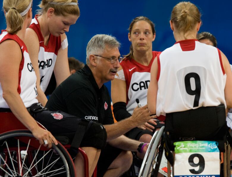 A group of four women in wheelchairs standing around a man wearing a black polo shirt kneeling speaking. 