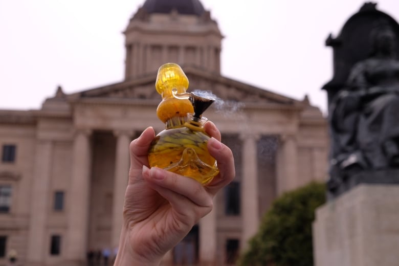 A hand holding a bong in front of the Manitoba Legislative Building.