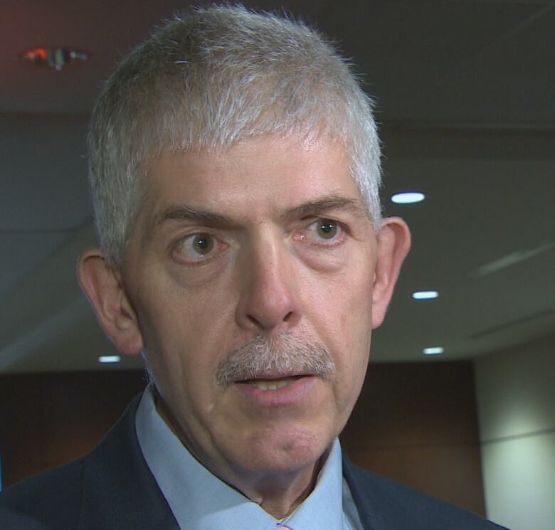 A man with short gray hair and a thin mustache looks slightly off to the right while facing the camera in a head and shoulders image. He's wearing a light blue dress shirt, and a dark blue suit. The background is an office building corridor or lobby.