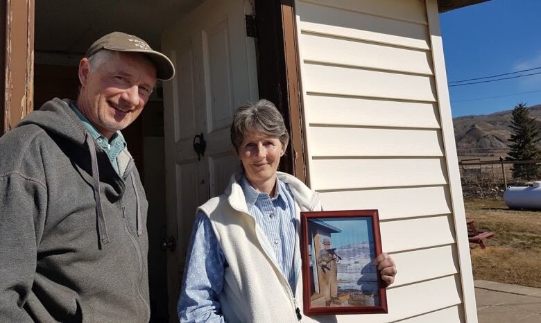 Two people stand at the door of their house, with one of them holding an old picture.