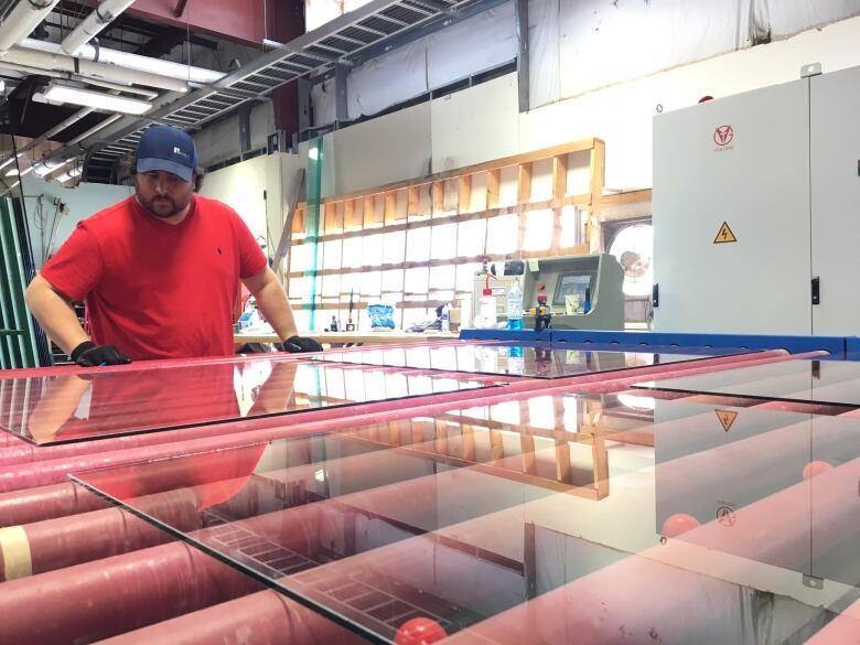 A bearded man in a red T-shirt, black gloves and black ball cap stands at a large piece of machinery holding a pane of glass.