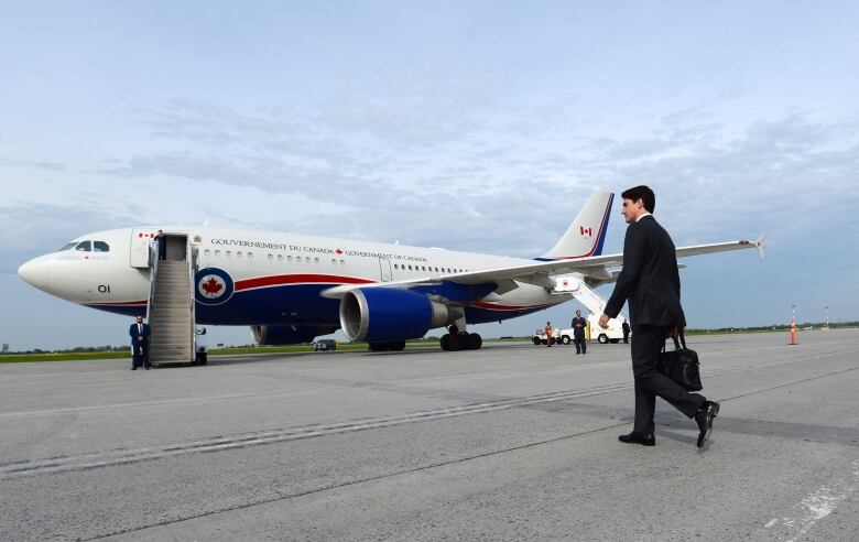 A man in a black wool coat walks towards a large airplane with a red maple leaf on the side on a tarmac on an overcast day.