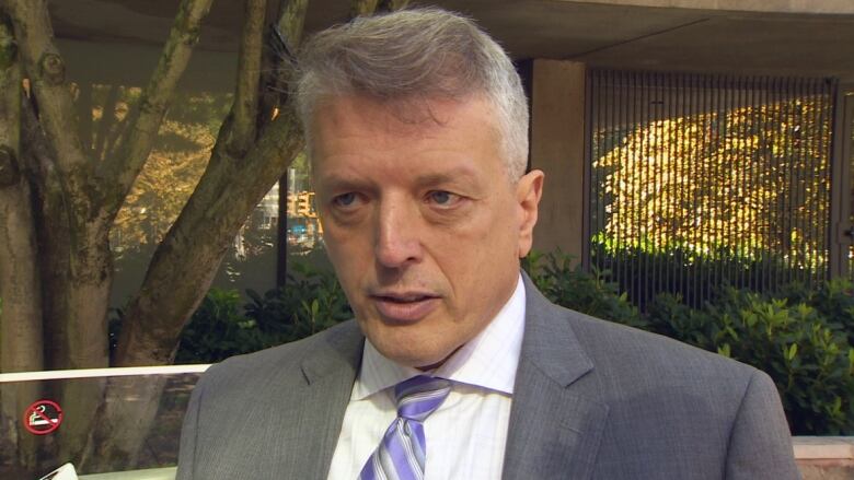 A man in a grey suit looks away from the camera in an outdoor environment.