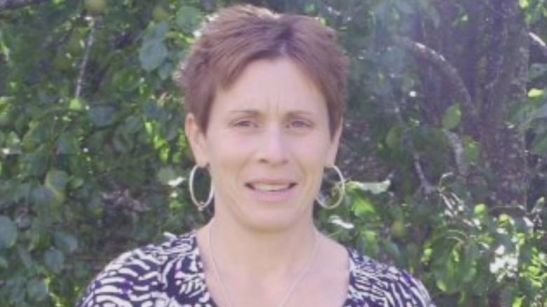 A woman with short brown hair and large hoop earrings is shown against a backdrop of trees.