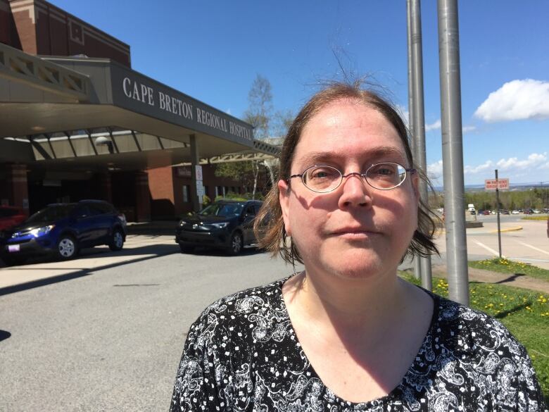 A woman with her hair pulled back, wearing glasses and a black-and-white print blouse, stands in front of the entrance to a hospital.