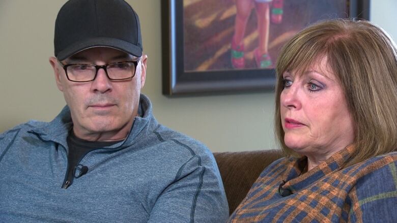 A man and a woman with serious expressions sit on a couch in a home.