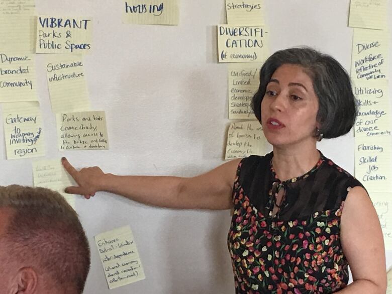 A woman points a white board covered in sticky notes.