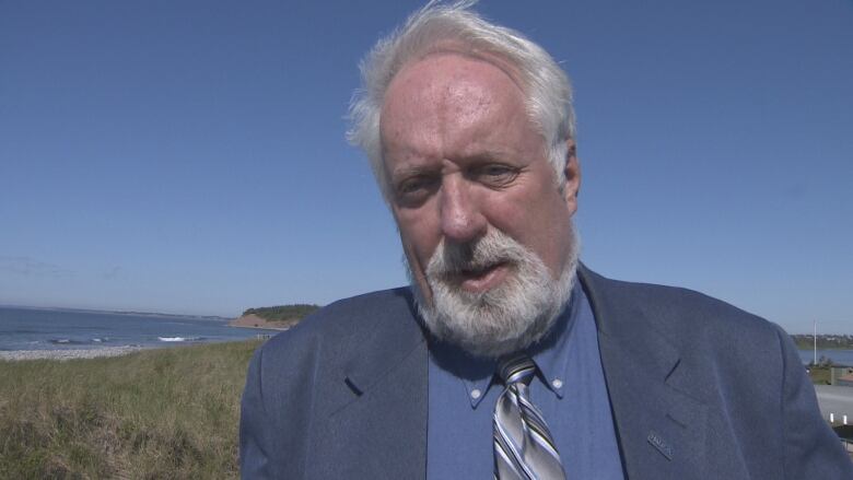 A white man with a blue shirt, navy jacket and tie stands outside in a coastal area. Blue water is visible behind him