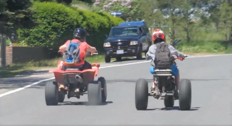 Two people on ATVs ride side by side on a residential road.