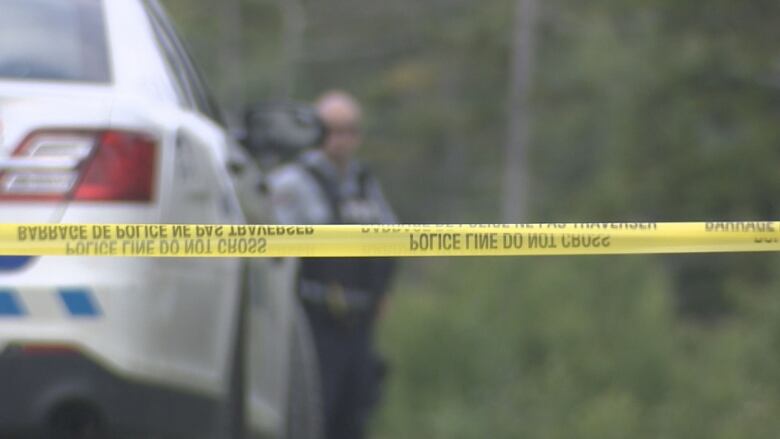 Yellow police tape is seen strung across a scene with an out-of focus RCMP cruiser and officer in the background