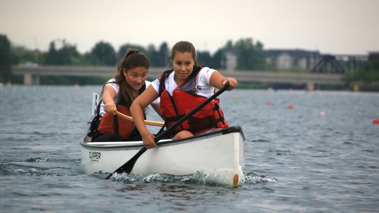 Two girls with brown hair canoeing on a lake. The canoe is white. They are wearing white t-shirts and red safety life jackets.