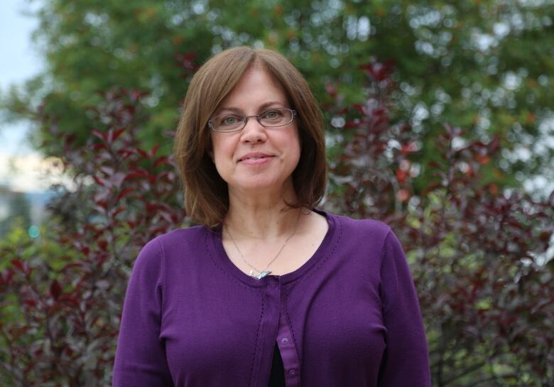 Woman with above shoulder length hair, wearing purple shirt, smiling at camera with a bush in the background.