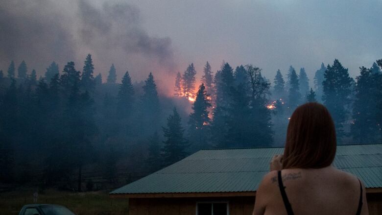 A woman looks out at a wildfire at dusk.