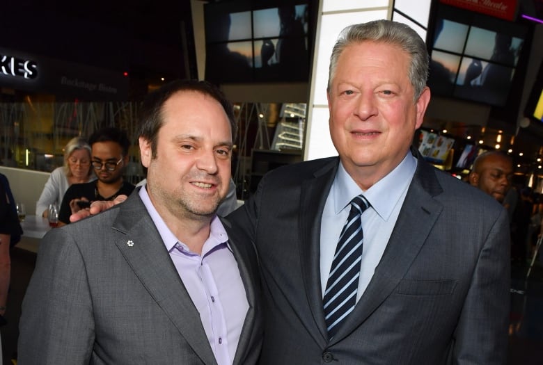 Two men in suits pose with their arms around one another.