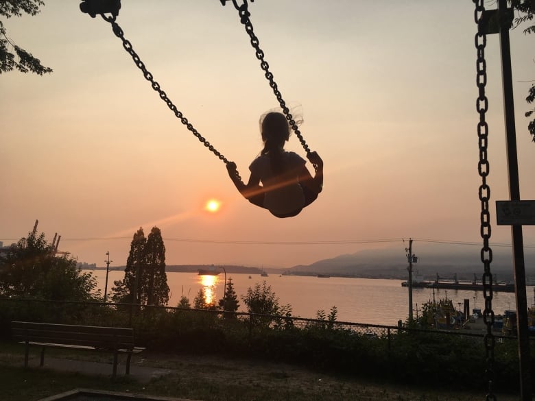 A silhouette of a young girl on a swing against a backdrop of Burrard Inlet with orange skies due to wildfire smoke and sunset.