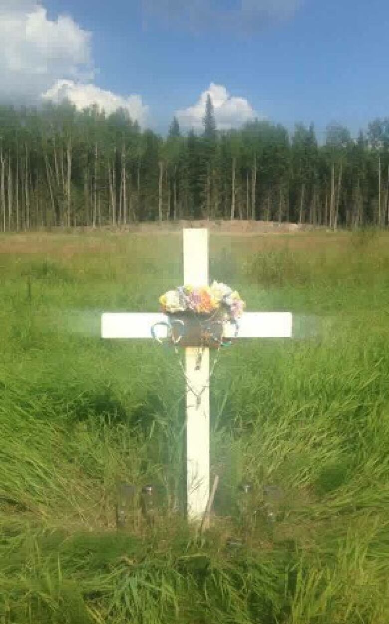 A white cross in a grassy area in bright sunshine, with a forest in the background.