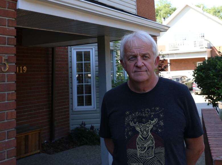 Terry Wickham in a black tee-shirt standing in front of a brick house. 