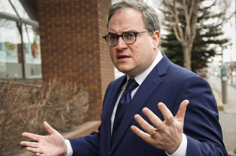 Man with glasses in blue suit gestures outside brown brick building.