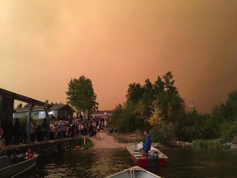 A crowd of people stand near a shoreline. A boat floats in the water.