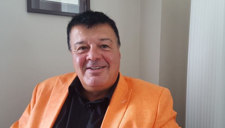 A man with dark hair, wearing a black shirt and orange jacket, sits and looks at the camera.