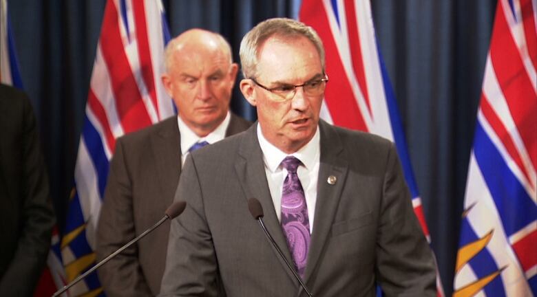 A white man speaks in front of a row of B.C. flags, with another white man behind him.