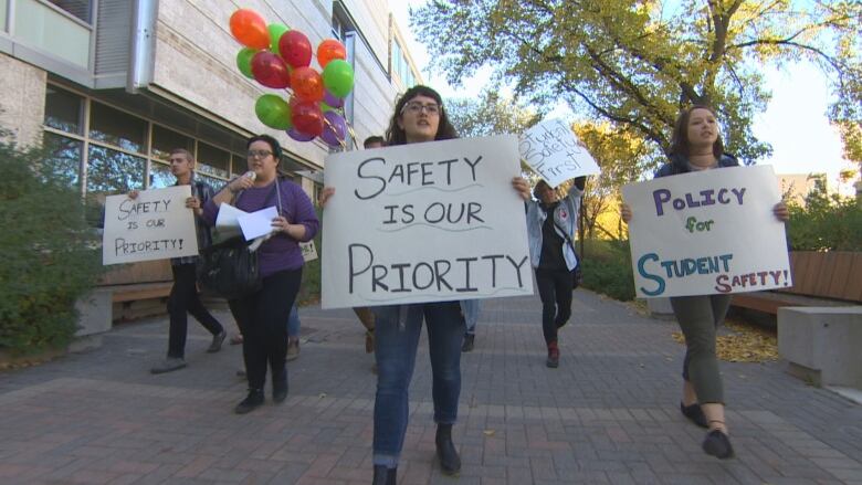 Students walk on a university campus carrying signs, with messages reading 