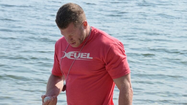 A muscular white man with short-cropped brown hair, wearing a red t-shirt and long black shorts is shown walking along a rocky shoreline. He is looking down at a stone in one of his hands.