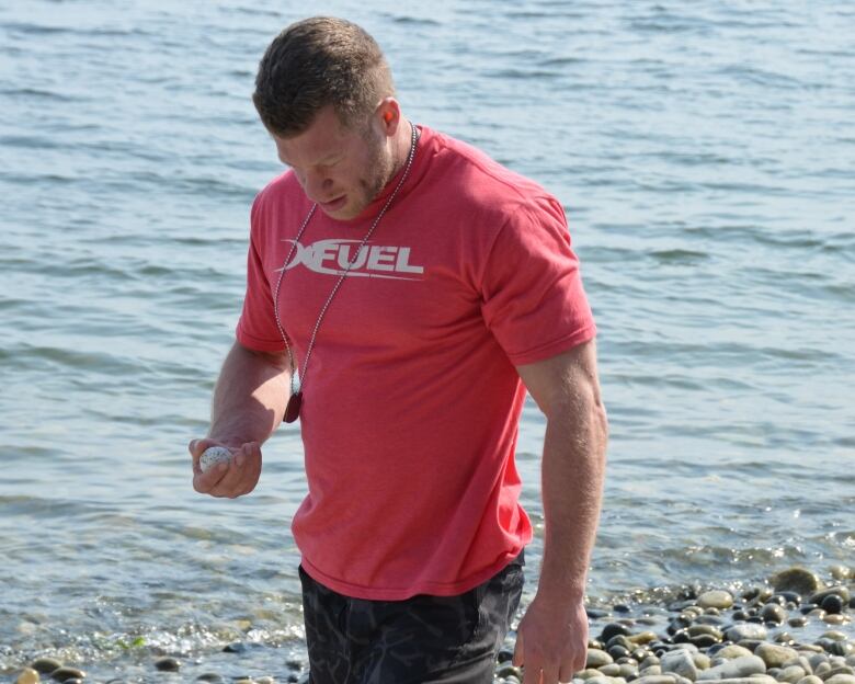 A muscular white man with short-cropped brown hair, wearing a red t-shirt and long black shorts is shown walking along a rocky shoreline. He is looking down at a stone in one of his hands.