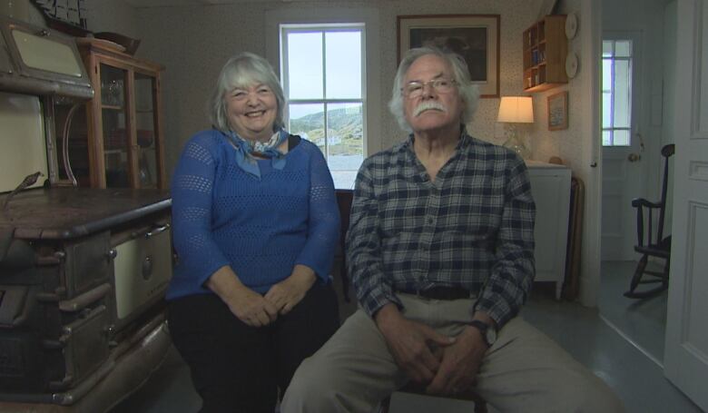 A smiling woman in a blue sweater and a man in glasses and plaid shirt sit next to each other in a seaside home.