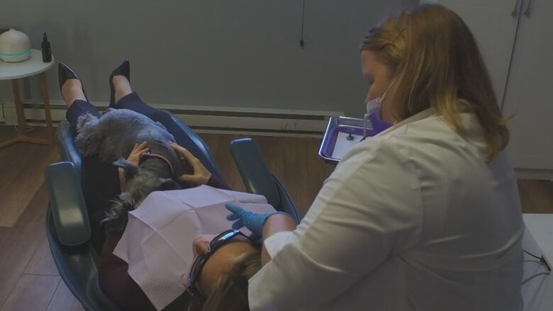 A woman in a white lab coat performs dental operations on a woman lying down. The patient has a dog on her lap. 