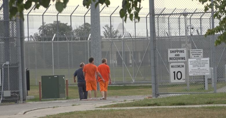 Two men in orange shorts and orange t-shirt walk beside a man in blue, in the shadow of large fences with barbed wire overtop. 