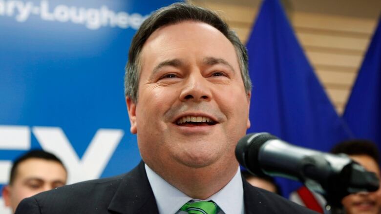 Jason Kenney smiles as he speaks into a microphone. He's wearing a dark suit and green tie.