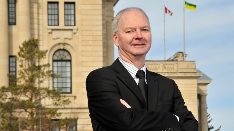 Man in black suit with his arms folded. There is a yellow-stoned building behind him.