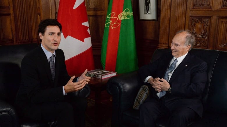 Two men in suits sit talking on a couch.