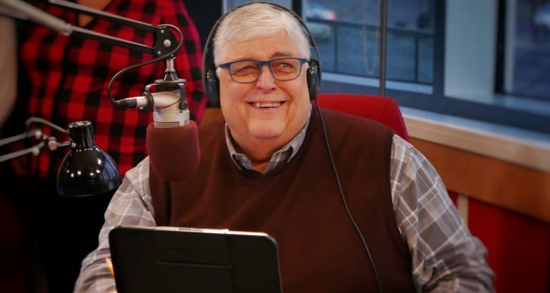 A white man with white hair smiles in front of a radio mic.