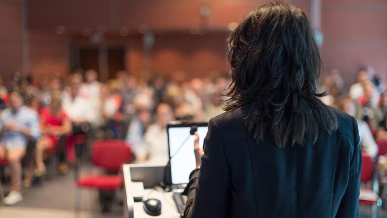 A lecturer is depicted from behind a podium speaking to a hall full of people listening in.