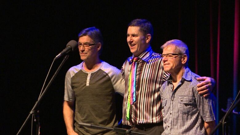 Three men stand smiling in front of a microphone on stage.