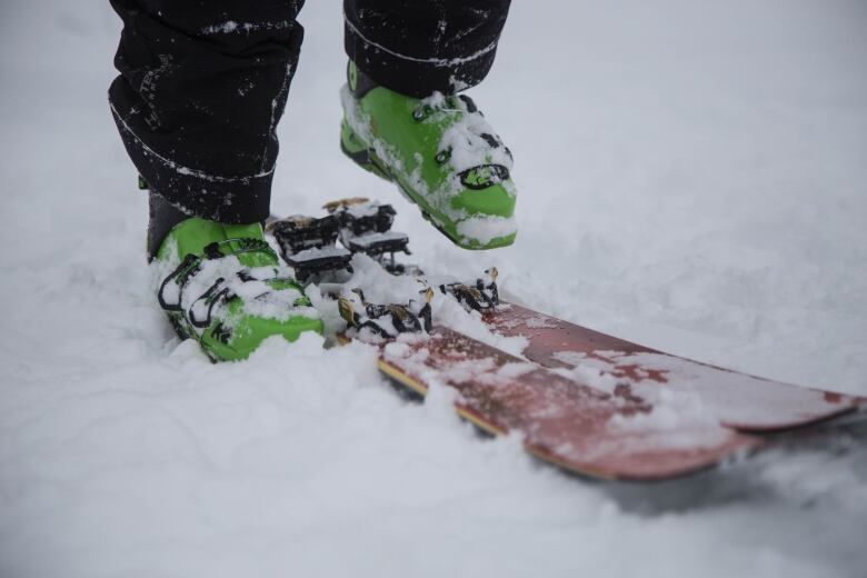 A close-up picture of a person with green ski boots clipping their feet into a pair of red skis in snow-covered ground.