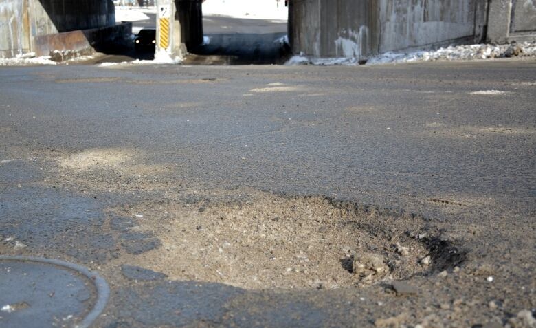 A large pothole is in the middle of College Street in Sudbury heading down towards a railroad underpass with crumbling concrete. 