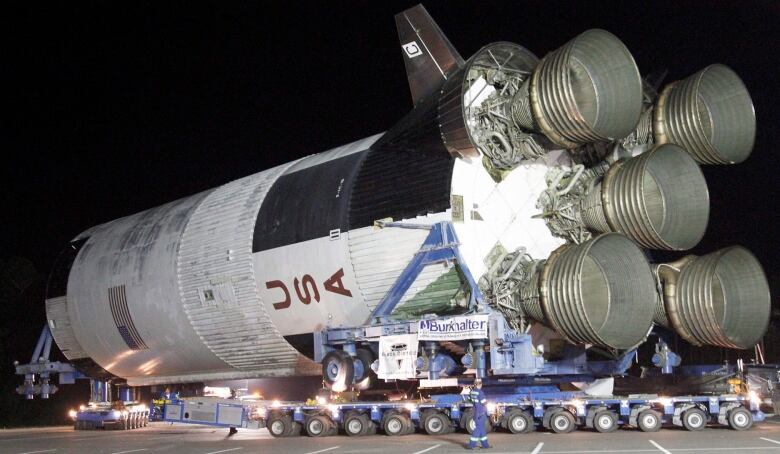 The Saturn V rocket is getting pulled in a parking lot on its side with five giant engines at its rear.