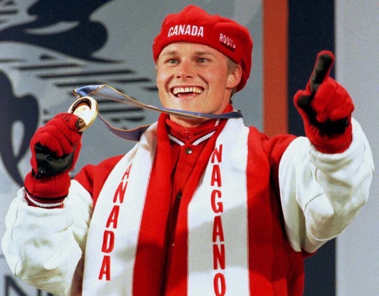 A smiling Olympic athlete dressed in Canadian red and white is shown wearing a gold medal around his neck, and holding the medal up in his right hand.