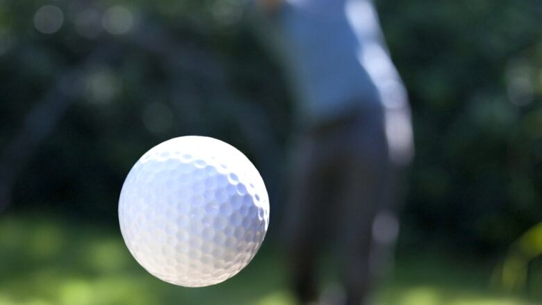 A white golf ball flies through the air with a blurry golfer swinging a club in the distance