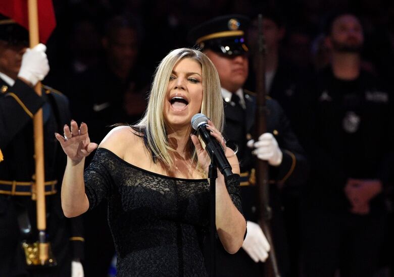 Singer Fergie performs the national anthem prior to an NBA All-Star game in black off the shoulder top, singing into microphone on stand.