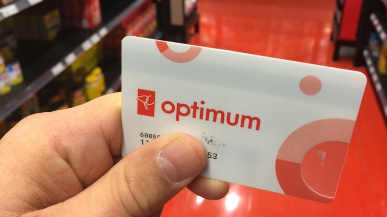 A loyalty card with the words PC Optimum on the front is seen close-up in a person's hand. The aisle of a retail store is seen in the background.