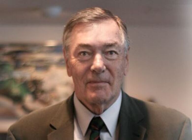 An older man wears a white collared shirt and brown suit jacket and smiles at the camera