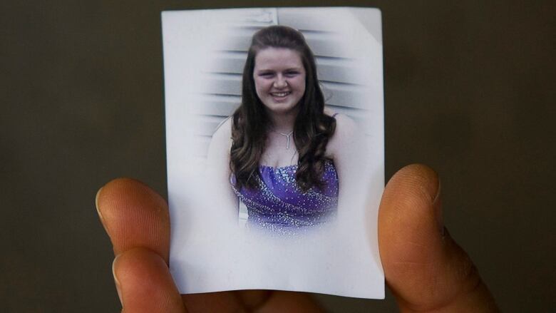 A hand is shown holding an image of a white woman in a sparkly purple dress with brown hair. The woman is smiling.